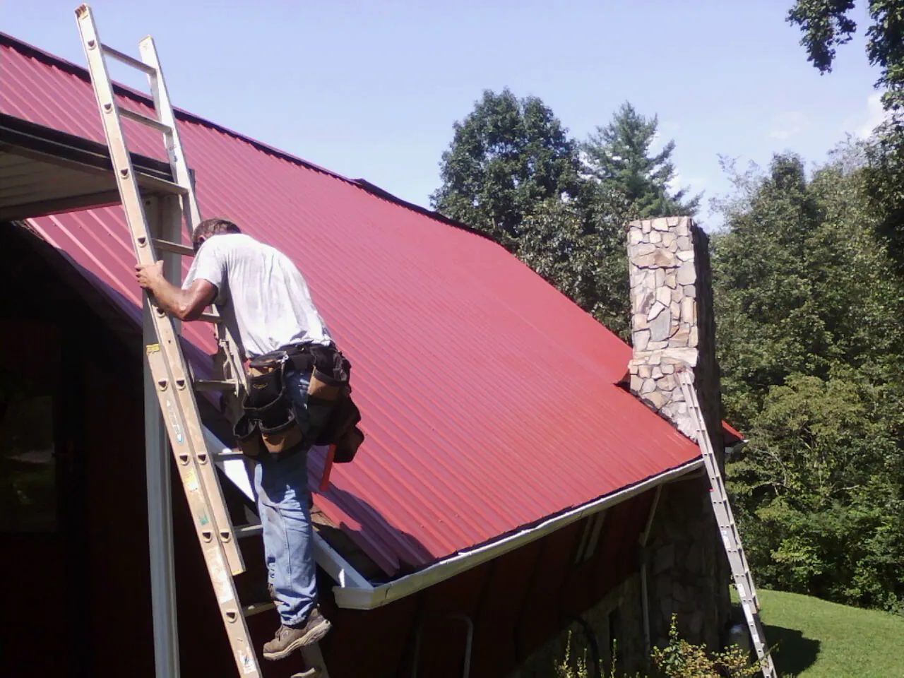Roofer on a ladder beside a red metal roof and stone chimney — Soffit & Fascia Repair by Roofing Brothers in Little Suamico