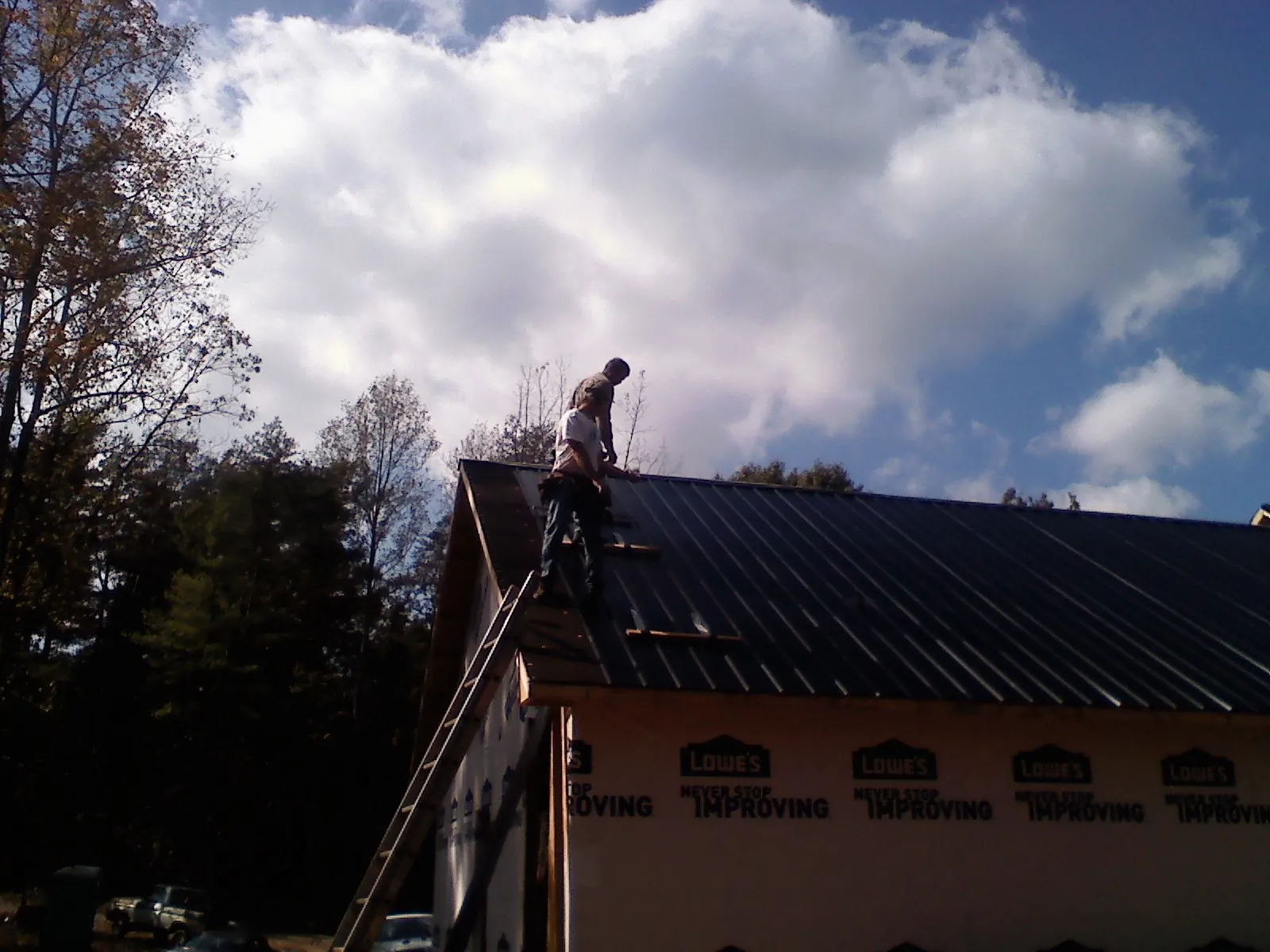Roofer fastening dark metal panels high against a cloudy sky, from a Roof Inspection job in Little Suamico